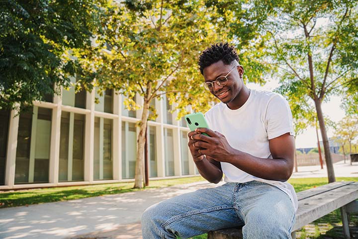 College student using his mobile phone sitting on a bench on a college campus